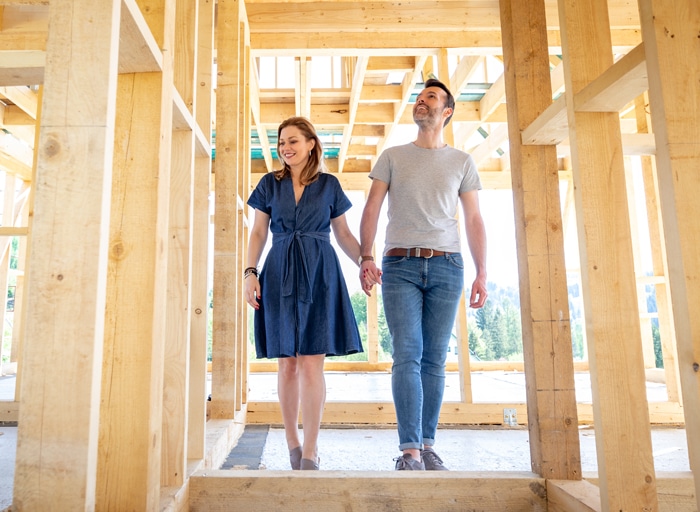 A man and a woman holding hands, walking through the wood framing of a new home construction.